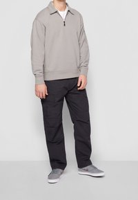 Sudadera gris claro con cuello de media cremallera, combinada con pantalones cargo oscuros que tienen múltiples bolsillos y zapatillas grises con acentos sombreados.