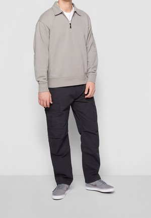Sudadera gris claro con cuello de media cremallera, combinada con pantalones cargo oscuros que tienen múltiples bolsillos y zapatillas grises con acentos sombreados.