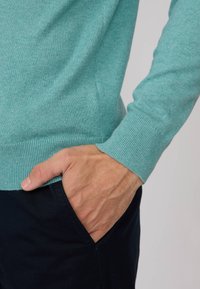 Un pull turquoise clair avec des bords côtelés au niveau des poignets et de l'ourlet, porté avec un pantalon marine foncé. Le tissu semble doux et texturé. Une main repose dans la poche.