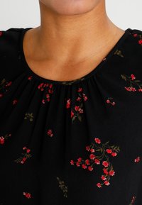 Svart topp med röda blommönster, plisserad halsringning och mjuk tygtextur. Accenter inkluderar gröna blad bland blommorna.