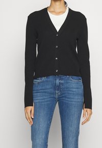 Kvinde iført en sort knappet cardigan over en hvid top og blå denimjeans, stående mod en ensfarvet lys baggrund.