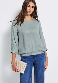 Blusa leggera di un verde chiaro, con scollo a barca, maniche a tre quarti e orlo arricciato, abbinata a jeans blu e una clutch intrecciata beige.