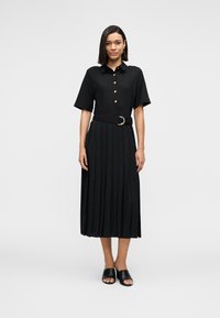 Μαύρο πλισέ φόρεμα με κοντά μανίκια, κουμπιά μπροστά και ζώνη με στρογγυλή αγκράφα. Το μοντέλο φοράει μαύρα σανδάλια με ανοιχτά δάχτυλα.