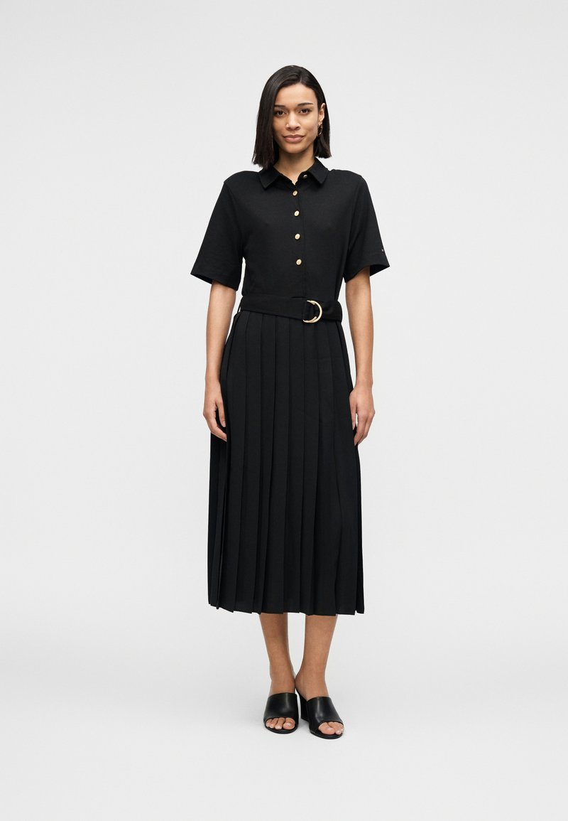 Μαύρο πλισέ φόρεμα με κοντά μανίκια, κουμπιά μπροστά και ζώνη με στρογγυλή αγκράφα. Το μοντέλο φοράει μαύρα σανδάλια με ανοιχτά δάχτυλα.