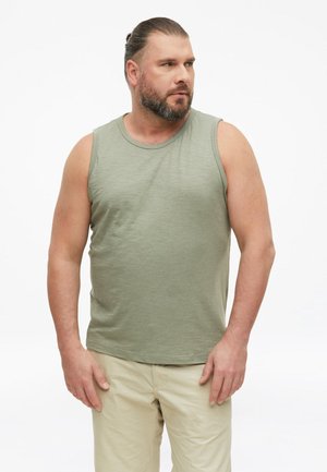 Mann mit Bart und zurückgebundenem Haar, der ein hellgrünes ärmelloses Shirt und beige Hose trägt, schaut seitlich vor weißem Hintergrund.