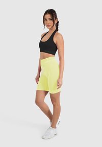 Zwarte sportbh met logo, gecombineerd met felgele high-waisted fietsbroek. Model draagt witte sportschoenen, heeft gevlochten haar, op een neutrale achtergrond.