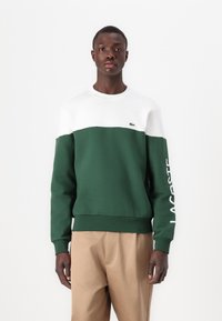 Sudadera color bloque en verde y blanco con detalles acanalados, con "LACOSTE" impreso en blanco en la manga derecha. Diseño liso, material de algodón.