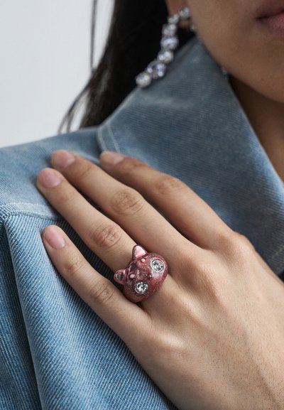 Roka z okrasnim prstanom v obliki rožnate živalske glave z velikimi prosojnimi dragimi kamni za očmi, ki počiva na rami modre jeans jakne.