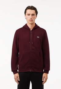 Felpa con zip di colore bordeaux in misto cotone, con una zip frontale, grandi tasche, cappuccio e un piccolo logo verde sul lato sinistro del petto.