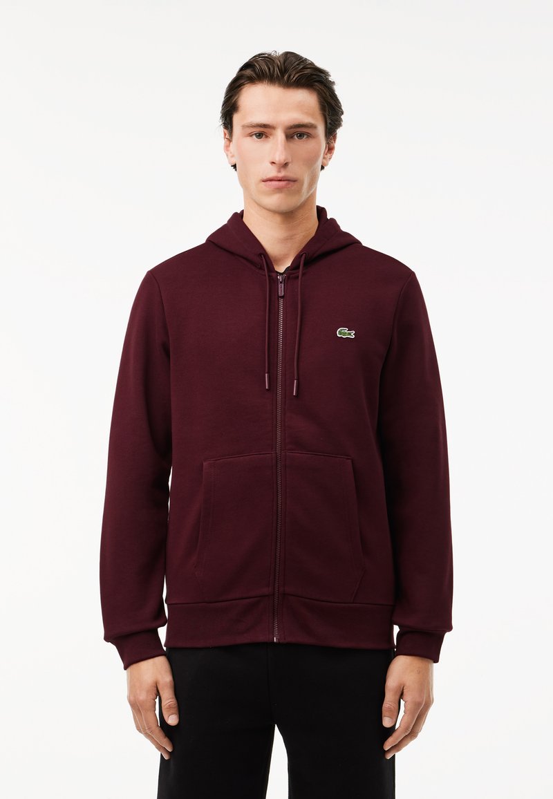 Felpa con zip di colore bordeaux in misto cotone, con una zip frontale, grandi tasche, cappuccio e un piccolo logo verde sul lato sinistro del petto.