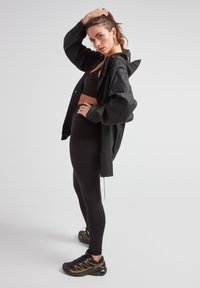 Svart vattentät jacka med huva, kombinerad med en svart sport-bh och högmidjade tights. Svarta träningsskor med gula accenter.