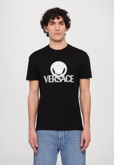 Fekete pamut póló, kerek nyakkivágással, rövid ujjakkal, elöl egy görög mintázattal és fehér „VERSACE” felirattal.