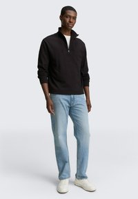 Zwarte rits-trui met geribde manchetten, gecombineerd met lichtblauwe jeans en witte sneakers. De figuur staat tegen een neutrale achtergrond.