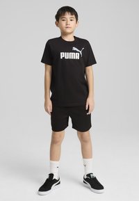 T-shirt en coton noir avec le logo « PUMA » blanc, associé à un short noir. Porte des baskets noires et des chaussettes blanches avec des accents de logo.