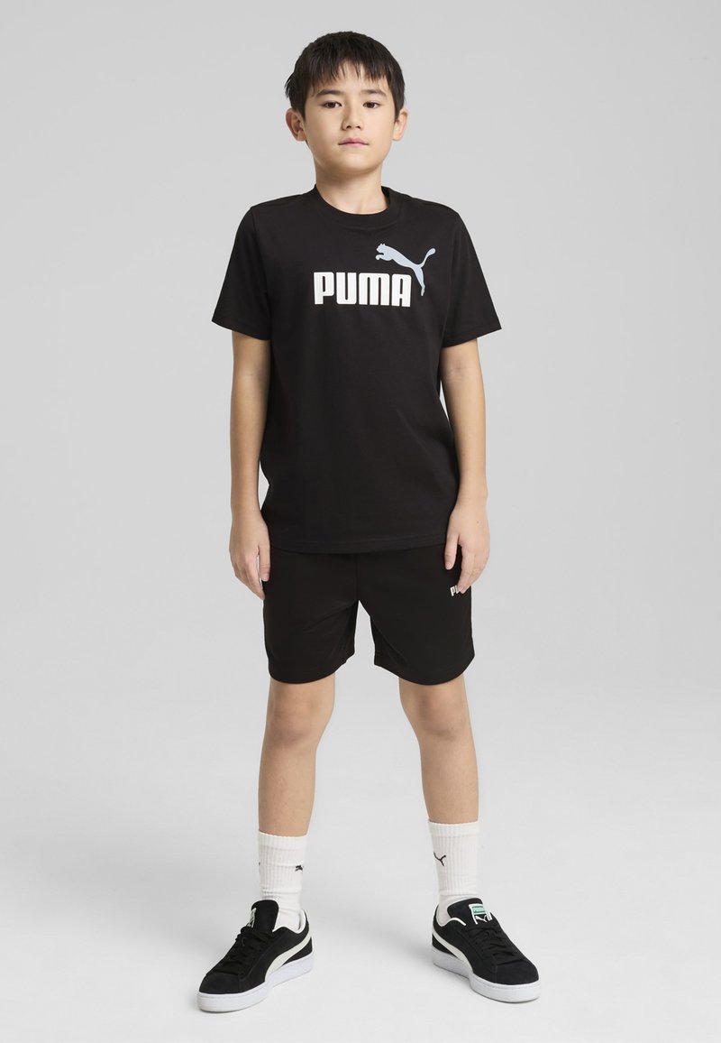 T-shirt en coton noir avec le logo « PUMA » blanc, associé à un short noir. Porte des baskets noires et des chaussettes blanches avec des accents de logo.