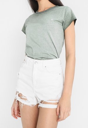 Woman wearing a green short-sleeve t-shirt tucked into high-waisted white distressed denim shorts with frayed edges.