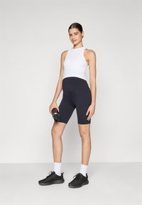 Top bianco senza maniche, pantaloncini sportivi neri e scarpe da ginnastica nere con calze bianche. Persona che tiene in mano una bottiglia d'acqua, in piedi davanti a uno sfondo neutro.