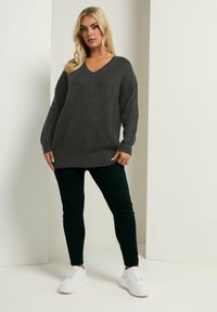 Dunkelgrauer gestrickter Pullover mit V-Ausschnitt, langen Ärmeln und lockerer Passform. Kombiniert mit dunkelgrünen Slim-Fit-Hosen und weißen Sneakers.