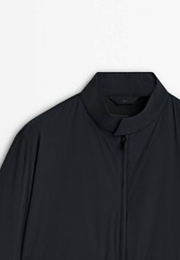 Veste noire avec un col court montant et une fermeture éclair frontale, affichée sur un fond clair uni.