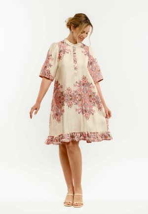 Kobieta ubrana w kremową sukienkę za kolano z różowo-pomarańczowymi kwiatowymi wzorami, z falbanką na dole i krótkim rękawem, stojąca i uśmiechająca się z opuszczonymi oczami.