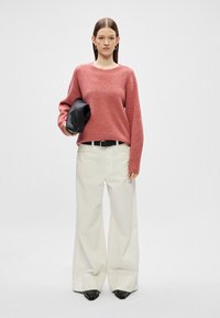 Roza pletena jopica z teksturiranim završkom, v kombinaciji z širokimi belimi hlačami in črnimi čevlji z ostrimi noski. Dodatki vključujejo črno torbico.