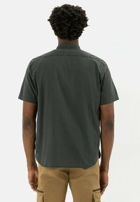 Camicia a maniche corte di colore verde scuro con colletto, orlo dritto e texture liscia, vista da dietro su uno sfondo neutro.