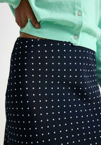 Cardigan verde menta con orlo a coste, abbinato a una gonna blu navy a pois bianchi. La gonna ha una texture liscia e un design aderente.