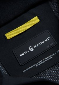 Svart tygetikett med "SAIL RACING"-logotyp i vitt, med en framträdande gul flik ovanför. Texten inkluderar "MEDIUM" och detaljer om design/fabrikation.