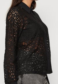 Black lace shirt with a button-down collar, featuring intricate cut-out patterns and long sleeves. Sheer fabric with a textured design.