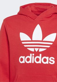 Röd huvtröja med vitt Adidas trefoil-logotyp och text centrerad på bröstet, med en framficka i kängeroustil och dragsko för huvan.