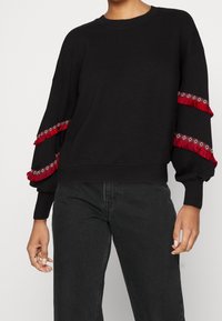 Svart sweatshirt med långa, puffiga ärmar, som har röda fransdetaljer och dekorativ sömnad längs manschetterna. Mjukt material, avslappnad passform.