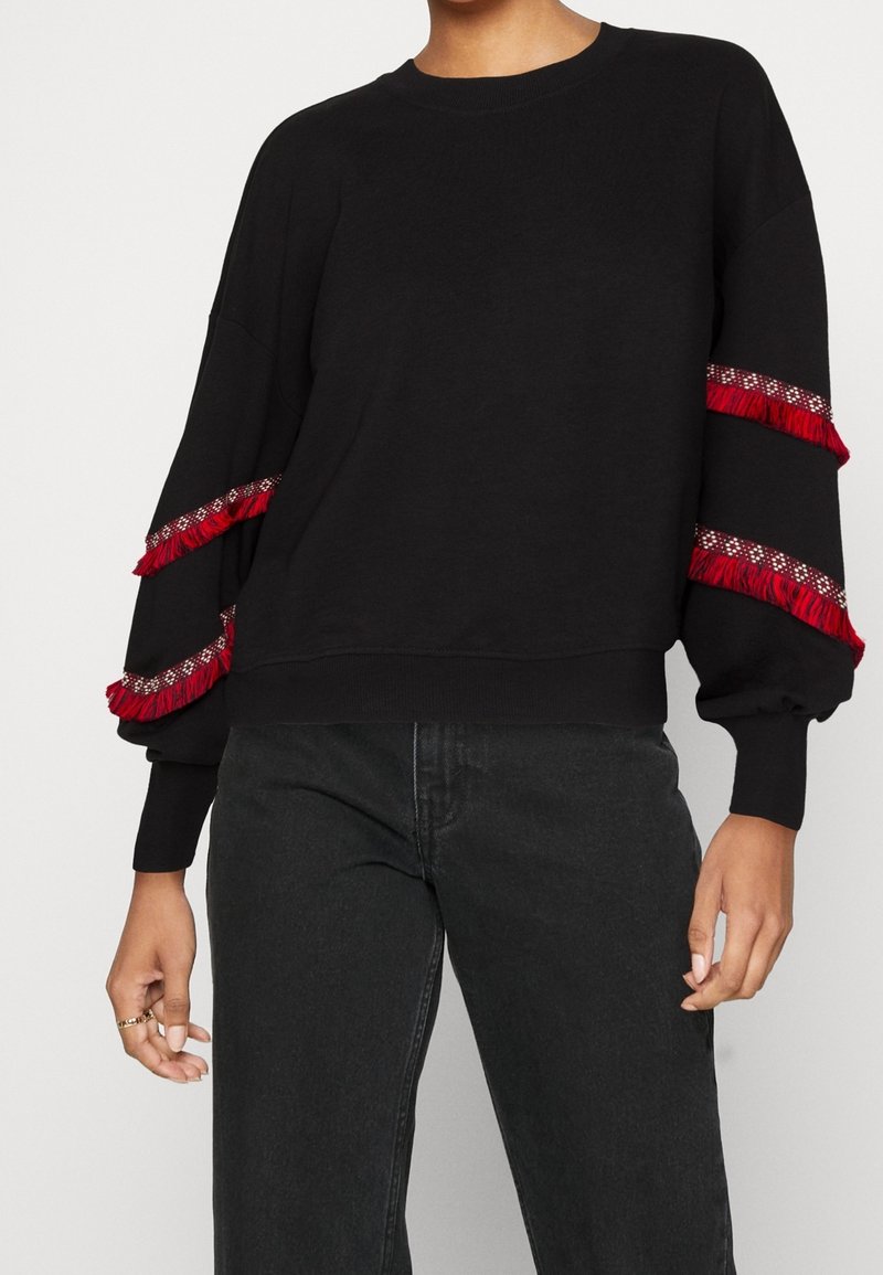 Svart sweatshirt med långa, puffiga ärmar, som har röda fransdetaljer och dekorativ sömnad längs manschetterna. Mjukt material, avslappnad passform.
