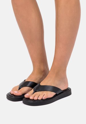 Füße in schwarzen Leder-Zehentrennern mit dicken Sohlen vor hellem Hintergrund, natürliche Haut und lackierte Zehennägel sichtbar.