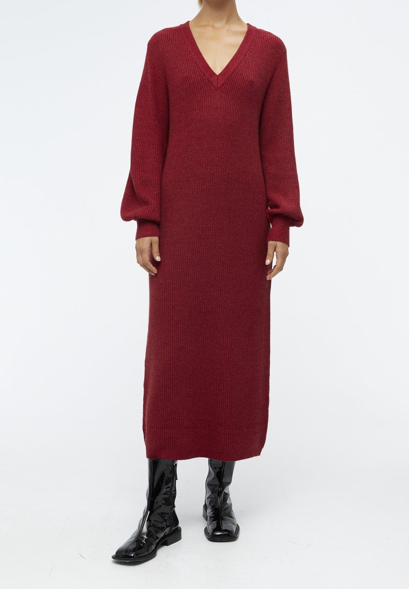 Femme portant une robe midi en maille rouge foncé à manches longues avec un col en V et des bottes mi-mollet en cuir verni noir, debout devant un fond blanc.