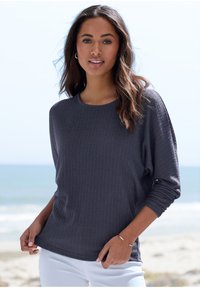 Eine Frau mit langen, welligen Haaren, die ein eng anliegendes, dunkel gestreiftes Langarmoberteil und weiße Hosen trägt, steht an einem Strand mit den Wellen des Ozeans im Hintergrund.