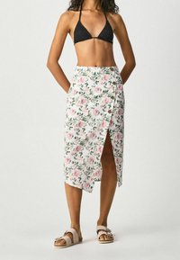 Jupe portefeuille florale avec une base blanche et des motifs de fleurs roses et vertes, fente latérale et boutons. Associée à un haut de bikini noir et des sandales.
