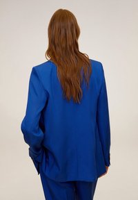 Blazer bleu sur mesure en tissu lisse, présentant une silhouette structurée, des épaules larges et une fermeture à un bouton.