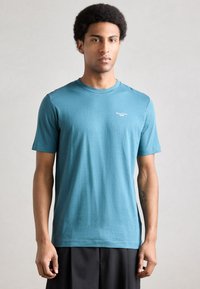 Tricou cu mâneci scurte, culoare teal, din țesătură moale. Are un decolteu rotund și un mic logo alb pe partea superioară stângă, purtat cu pantaloni negri.