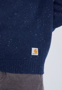 Navy gebreide trui met gespikkelde textuur, geribbeldezoom en een klein Carhartt-logo in oranje en wit onderaan aan de linkerkant.