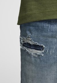 Näruppskott av blå denimjeans med en horisontell reva som avslöjar mörkt tyg under, kombinerat med en olivgrön skjorta.