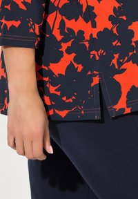 Maglietta a maniche corte rossa con un motivo floreale blu scuro, dotata di una fessura laterale e cuciture a contrasto visibili. Tessuto liscio e elasticizzato.