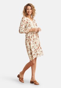 Kurzarmkleid in Creme mit braunem abstraktem Muster und V-Ausschnitt. Getragen mit braunen Blockabsatzsandalen.