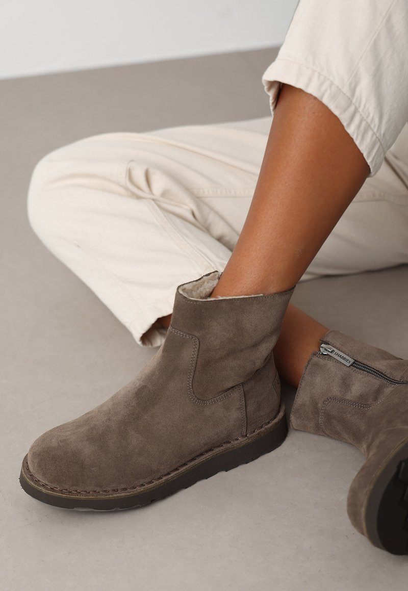 Botas de tobillo de ante marrón con un tacón bajo, cierre de cremallera y forro suave, sobre un fondo neutro. Estilo casual con un aspecto ligeramente desgastado.