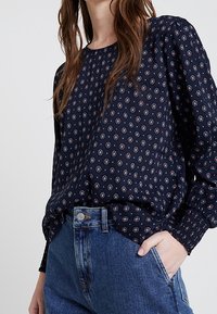 Blusa a maniche lunghe blu navy con motivo a rombi in rosso e bianco. Tessuto morbido, vestibilità comoda e polsini arricciati. Abbinata a jeans a vita alta.