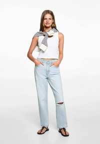 Top bianco corto, jeans a vita alta blu chiaro con dettagli strappati, sandaletti neri e un maglione a righe bianche e nere appoggiato.