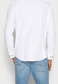 Valkoinen pitkähihaista paitaa, jossa on napilliset hihansuut, sileä kangas, kaareva helma ja mustat yksityiskohdat hihansuissa, yhdistettynä vaaleansinisiin farkkuihin.