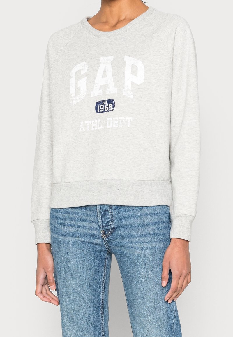 Sweat-shirt gris avec un col rond et des manches raglan. Arbore un logo "GAP" blanc et "ATHL DEPT" en impression usée. Associé à un jean bleu clair.
