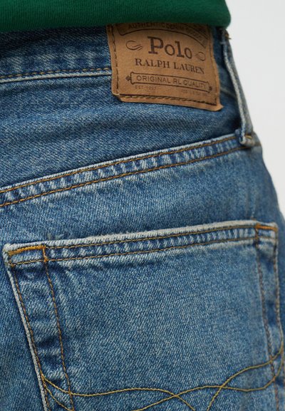 Κοντινό πλάνο της πίσω τσέπης από μπλε τζιν με καφέ δερμάτινο λογότυπο της Polo Ralph Lauren και πορτοκαλί ραφές.