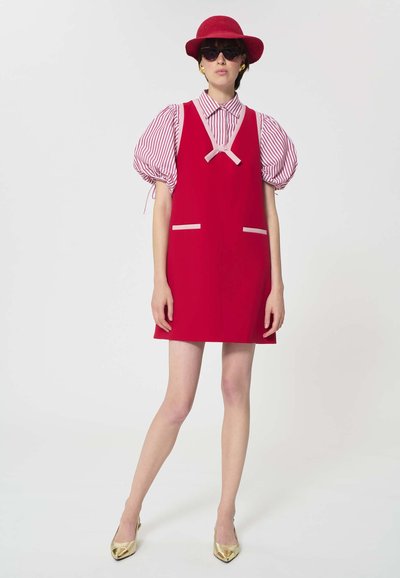 Femme portant une robe rouge sans manches sur une chemise à manches bouffantes rayée rose et blanche, un chapeau rouge, des lunettes de soleil noires et des chaussures pointues dorées.