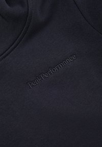 Tessuto in pile blu scuro con logo "PeakPerformance" a rilievo. La superficie liscia e il design minimalista accentuano la sua semplicità.